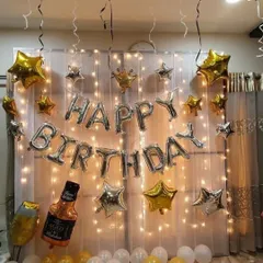 For this birthday setup, I used a wall of fairy lights behind a sheer white curtain to create a magical effect. The silver 'Happy Birthday' balloons are accented with gold and silver star balloons and a fun whiskey bottle prop.