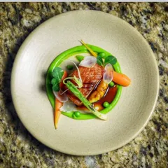 A beautifully plated main course. This dish features pan-seared duck breast on a bed of vibrant green pea puree, garnished with baby carrots, asparagus, and edible flowers for a fine-dining look.