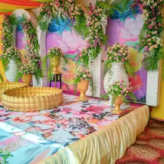 A colorful Haldi stage with a floral printed backdrop, golden lotus-shaped bowls (urli), and arrangements of pink and yellow flowers.