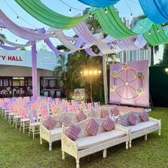 A whimsical outdoor Mehendi or Haldi setup featuring pastel-colored drapes, traditional carved white seating, and a unique Ferris wheel backdrop.