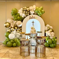 This First Holy Communion stage decoration uses a green, white, and gold color scheme. The setup includes an arched backdrop with a cross, a balloon garland, and stylish dessert stands.