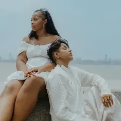 A serene and contemplative portrait of the couple sitting by the sea, with the Mumbai skyline in the distance. A quiet moment of togetherness.