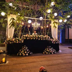 An elegant outdoor bar setup perfect for a sophisticated garden party. The bar is heavily decorated with white and pink flowers, set under a canopy of bamboo with hanging lanterns and a chandelier.
