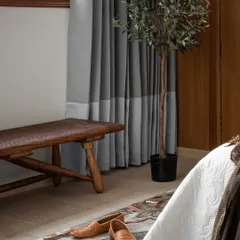 A detail shot showing the interplay of textures: a floral rug, a rustic wooden bench, and two-toned grey curtains. These elements add depth and character to the serene bedroom environment.
