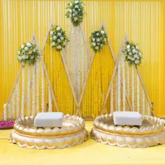 Here is the main stage for the Haldi ritual, featuring two traditional bajots. The backdrop is a geometric wooden frame decorated with marigold and tuberose strings, blending modern design with traditional flowers.