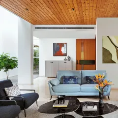 The living room of the Split House combines a warm teak wood ceiling with clean white walls and marble floors. The furniture and art add subtle pops of color, creating a balanced and sophisticated interior design.