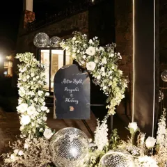 A "Retro Night" themed entrance for a Sangeet party. The decor includes a custom welcome sign, disco balls, and an arch of white flowers, setting a fun and nostalgic tone for the evening.