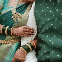 A detailed shot focusing on the bride's hands, adorned with intricate henna and traditional green bangles, resting on the groom's arm. It highlights the fine details of the saree border, kamarbandh, and the texture of the groom's jacket.