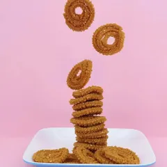 A creative and dynamic shot of chakli snacks appearing to fall and stack on a plate against a solid pink background, perfect for engaging social media content.