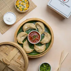 A serving of our classic steamed vegetable momos, presented on a banana leaf inside a steamer basket. They are served with a side of spicy red chutney and creamy mayonnaise.
