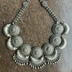 An antique-style oxidised silver necklace. This piece features a series of circular and crescent-shaped motifs with ghungroo drops, creating a substantial and eye-catching choker.