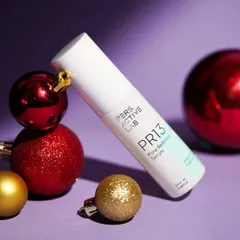A clean, festive product shot of a pore refining serum placed among shiny red and gold Christmas ornaments. The purple background provides a rich contrast, making the product pop.