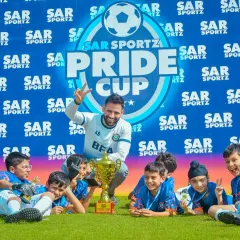 A coach and his team of young players celebrate with their trophy in front of the Sar Sportz Pride Cup banner.