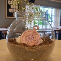 A charming nautical-themed table centerpiece. A glass bowl is filled with sand and a compass cutout, with delicate baby's breath flowers adding a soft touch.