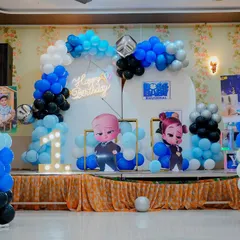 A Boss Baby themed first birthday party setup on a stage. It includes character cutouts of Boss Baby, a neon "Happy Birthday" sign, and a bold balloon arch in blue, black, and white.