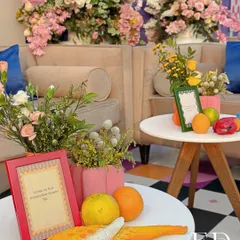 A vibrant seating area from the 'Elrow Carnival' event, featuring colorful floral arrangements, fresh oranges, and a whimsical parrot figurine. The checkered floor and pink accents add to the playful, high-energy atmosphere.