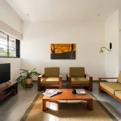 The living room is arranged with simple, modern wooden furniture. Large windows ensure the space is filled with natural light, while indoor plants help to blur the boundary between inside and out.