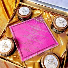 A vibrant and joyful invitation hamper. The bright pink wedding card is the star, surrounded by four glass jars filled with goodies, each topped with a custom monogrammed lid featuring the couple's initials.