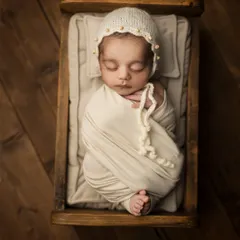 A sweet new blossom of humanity. This classic overhead shot shows a peacefully sleeping newborn, wrapped in neutral tones and wearing a delicate pearl-adorned bonnet in a tiny wooden bed.