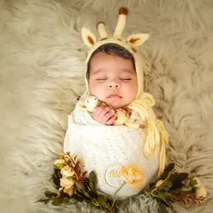 A sleeping newborn wearing an adorable giraffe-themed bonnet, swaddled in a cream-colored wrap on a soft, furry surface.