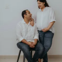 A full-length portrait showing the couple's relaxed dynamic during their home photoshoot. The simple posing against a plain wall keeps the focus entirely on their connection and anticipation.