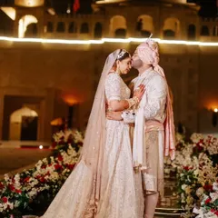 An intimate moment for the couple against the illuminated facade of a Jaipur palace. The wedding aisle is lined with lush red and white florals, creating a romantic pathway for their grand entrance.