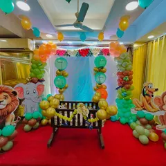 A playful jungle themed naming ceremony setup. This decor features a central cradle decorated with garlands, surrounded by a colorful balloon arch and life sized cutouts of cartoon animals like a lion, elephant, and monkey.