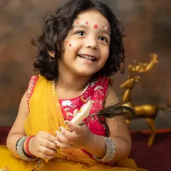 A beautiful little girl dressed as Radha for a Janmashtami themed shoot, holding a peacock feather with a joyful smile.