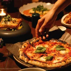 An intimate dinner scene, where a hand reaches for a slice of our freshly baked artisanal pizza, topped with basil and mozzarella.