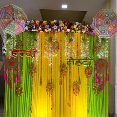 A detailed Haldi and Mehendi backdrop. This setup features bright yellow and green drapes, hanging floral strings, traditional Indian decor pieces, and embroidered umbrellas.