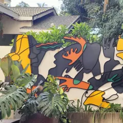 This colorful outdoor wall installation is made of layered, painted metal. The abstract, biomorphic shapes are designed to interact with the surrounding foliage, bringing a sense of playful energy and 'fun and frolic' to the exterior space.