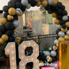 An 18th birthday celebration on a rooftop terrace. The marquee numbers are the centerpiece, surrounded by a black, gold, and silver balloon arch.