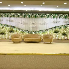 This elegant reception stage uses a classic white and green color scheme. The backdrop is made of white drapes and a curtain of string lights, with chandeliers and lush white floral arrangements adding layers of texture and light. It’s a timeless look that exudes sophistication.