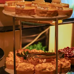 A two-tiered stand laden with our patisserie creations. The top tier holds delicate lemon meringue tarts, while the bottom features rustic apple crumbles, showcasing the range of our dessert offerings.