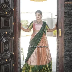 This peach and green silk half saree creates a beautiful silhouette, captured here in a doorway. It's a classic choice for weddings and festive occasions.