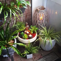 This tranquil corner features a miniature zen garden with a Buddha statue and floating candles. The arrangement is surrounded by various potted plants and a woven lantern, creating a perfect spot for meditation and peace.