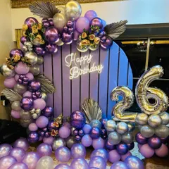 A modern purple paneled backdrop decorated with a lush arrangement of purple and silver balloons and dried palm leaves for a 26th birthday.