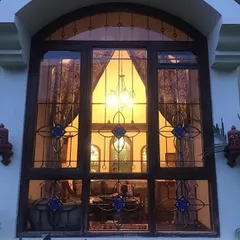 Seen from the outside at night, the large arched window of the Roopa Hariprasad Residence glows like a lantern. The intricate pattern of the custom grille is silhouetted against the warm interior light, creating a dramatic architectural feature.