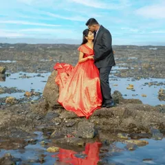 A beautiful pre-wedding portrait with a dramatic sky and water reflection. The bride's vibrant red gown contrasts beautifully with the serene landscape.