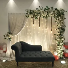 An elegant and simple engagement setup perfect for home functions. This features an L-shaped white floral arch, sheer curtains with fairy lights, and a comfortable chaise lounge for the couple.