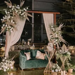For a modern and chic reception, we designed this sophisticated lounge area. The setup features a plush emerald green sofa, complemented by dried pampas grass and white floral arrangements on geometric gold stands, creating a look of understated luxury.
