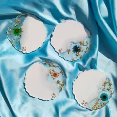 A set of four personalized resin coasters. The design combines a solid white base with a translucent section filled with gold flakes and a variety of colorful preserved flowers, each with a custom initial.