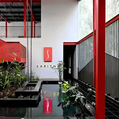 This wide shot of the SARITHA SERENE office highlights the interplay of industrial and natural elements. The exposed red steel frame and open upper level create a sense of volume, while the water feature and greenery ground the space.