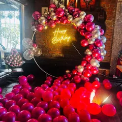 An indoor birthday party setup featuring a large ring backdrop. The circular frame is decorated with a dense arch of red and silver balloons and a central 'Happy Birthday' neon sign, with extra balloons covering the floor for a fun, immersive effect.