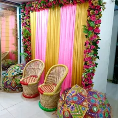 A vibrant photo corner for a Mehandi or Haldi event. I used pink and yellow drapes, a floral border, and traditional mooda chairs with colorful umbrellas to create this spot.