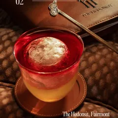A beautifully layered cocktail from The Hedonist. The lighting perfectly captures the separation of colors and the large, clear ice sphere. The branded menu in the background adds context and reinforces the bar's identity.