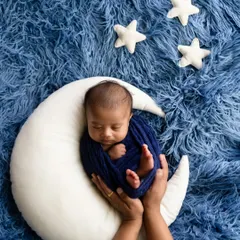 A newborn wrapped in deep blue, held in a crescent moon prop against a furry blue backdrop with soft white stars. This shot is a beautiful example of a celestial-themed photoshoot.