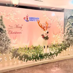 A festive 'Merry Christmas' photo backdrop for Goregaon Sports Club. The winter wonderland scene includes flocked trees, a decorative reindeer, and a white picket fence, creating a perfect holiday photo opportunity.