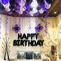 A birthday decoration at home with a purple, black, and white color theme. We filled the ceiling with balloons and hanging ribbons and placed a 'HAPPY BIRTHDAY' foil balloon set above the main seating area.