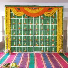 A traditional Haldi backdrop with a green base, decorated with hanging flower strings and a large toran of orange and yellow marigolds.